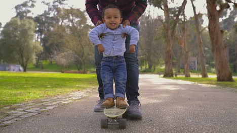Little-boy-standing-on-skateboard,-rolling-with-help-of-father